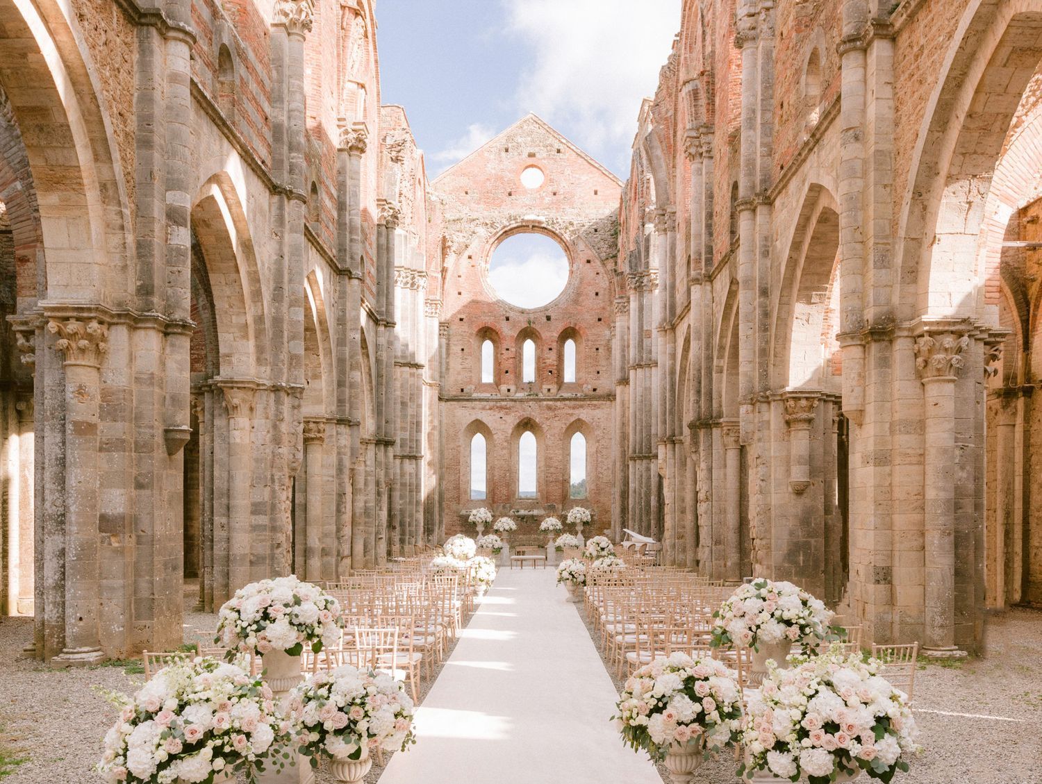 Elegant outdoor wedding ceremony setup inside the roofless ruins of San Galgano Abbey, adorned with white floral arrangements.