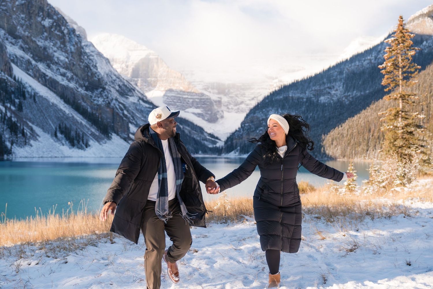Couple holding hands and running playfully at snowy Lake Louise with turquoise water and mountains in background in fall season.