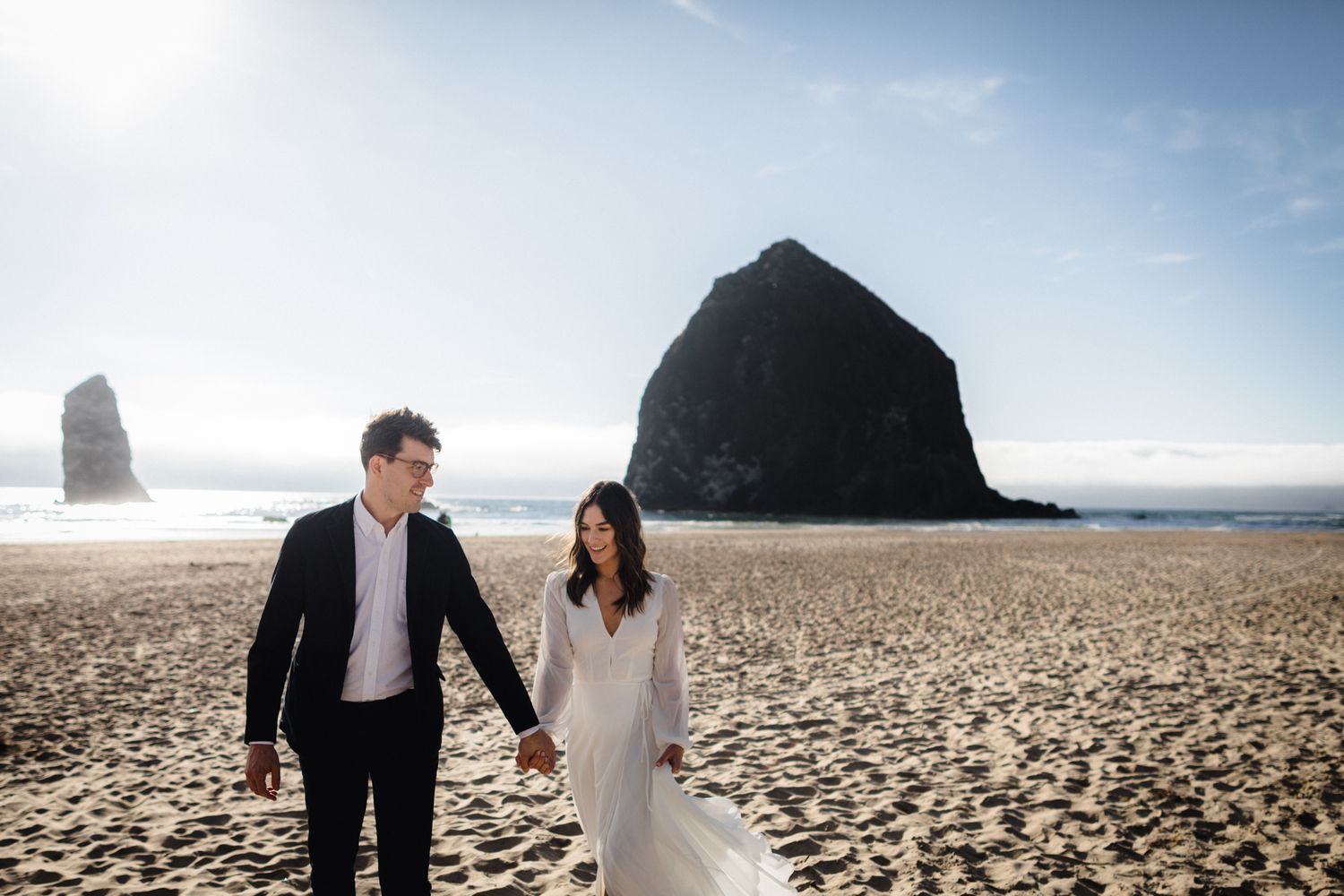 Couple in wedding attire holding hands on sandy beach with iconic Haystack Rock at Cannon Beach, Oregon.