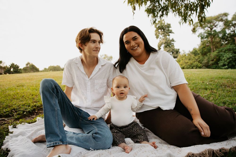 Family of three sitting on a blanket in a park, two adults in white shirts with a baby between them.