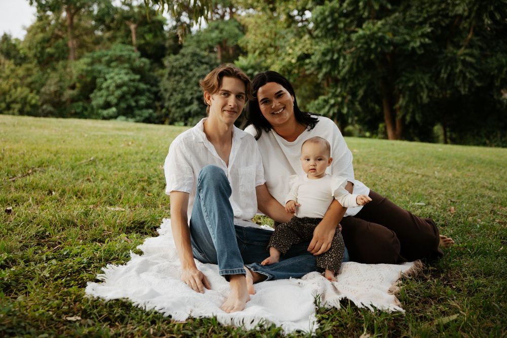 Young family with baby sitting on white blanket in lush green park, smiling at camera on sunny day.