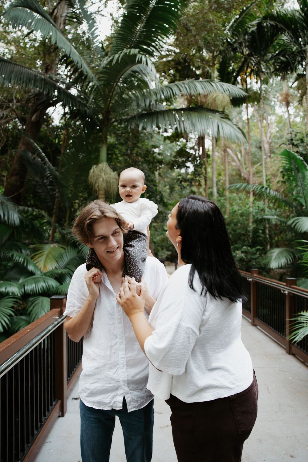 Two parents holding their baby on a wooden bridge surrounded by lush tropical greenery.