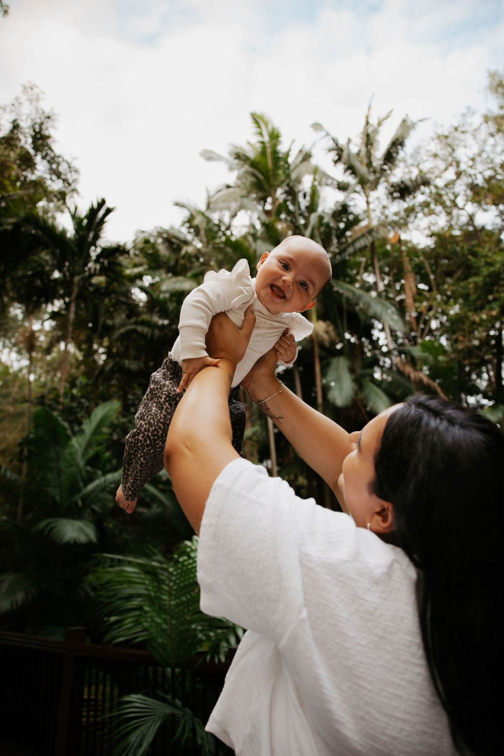 A mother joyfully lifts her smiling baby up in the air outdoors, surrounded by lush tropical greenery.