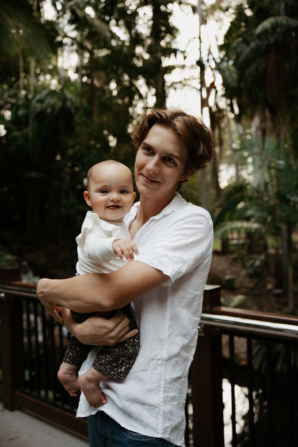 Young man in white shirt holding a smiling baby on a wooden deck surrounded by lush green trees.