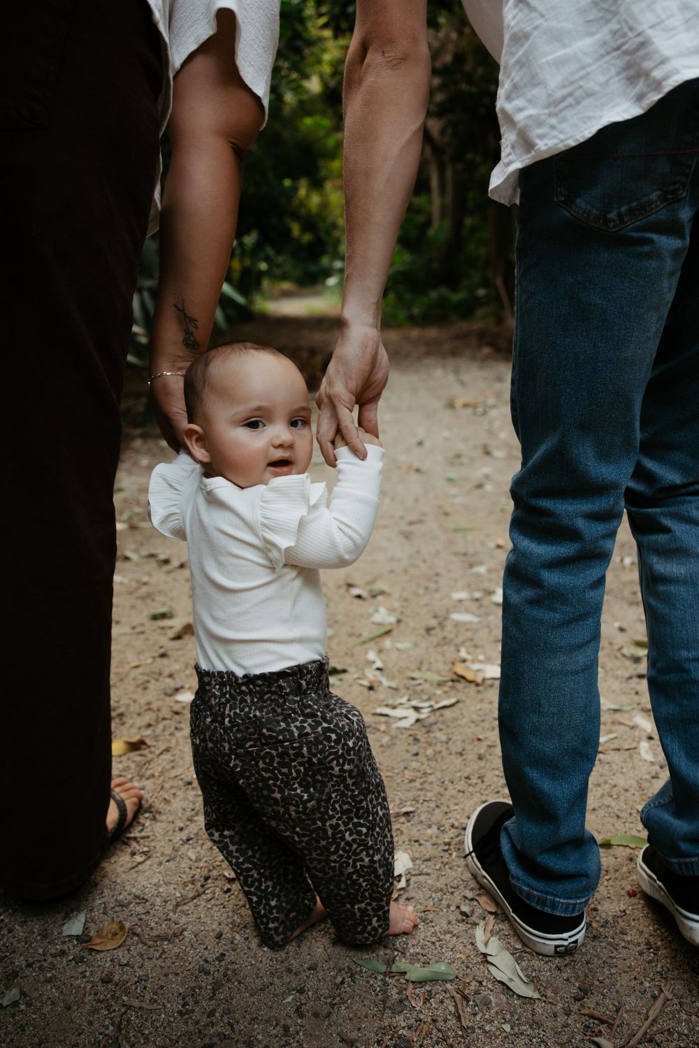 Baby taking first steps while being supported by parents hands outdoors.