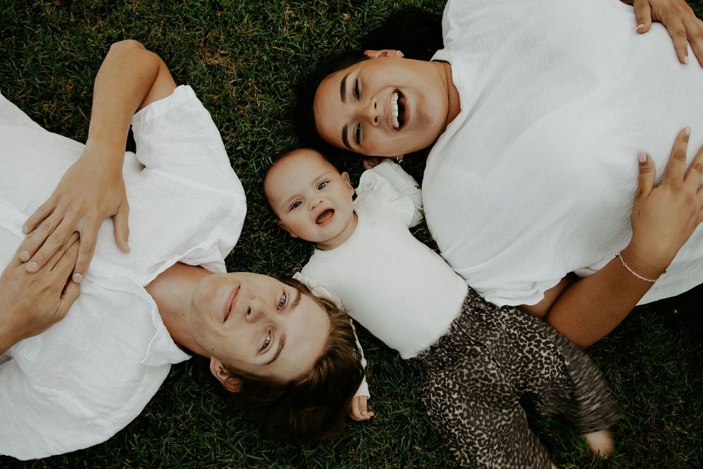 Two parents and a happy baby lying on green grass, viewed from above, all wearing white shirts.