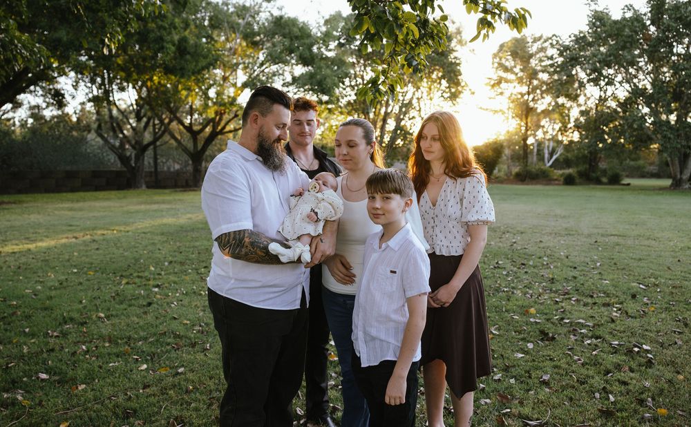 Family of five posing outdoors at sunset in a lush green park, with parents holding a baby and children nearby.