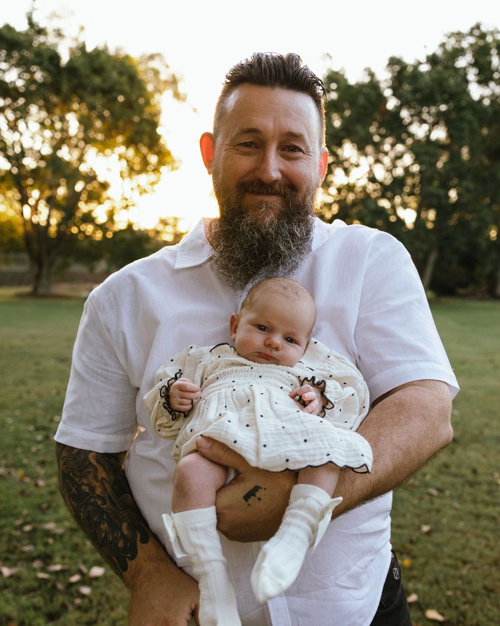 Bearded tattooed man smiling while holding a baby in a white outfit outdoors in a park at golden hour.