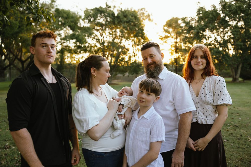 Family of six posing outdoors at golden hour, with parents holding a newborn baby surrounded by lush green trees.