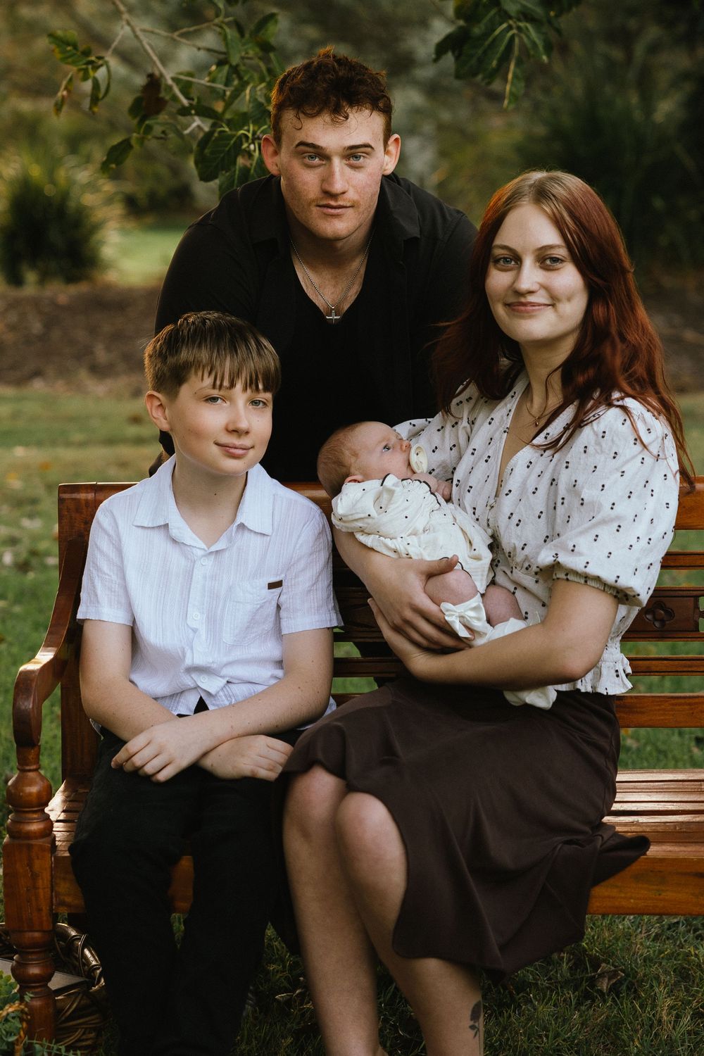 Family portrait on a bench outdoors, parents with a newborn baby and young boy, smiling in a park setting.