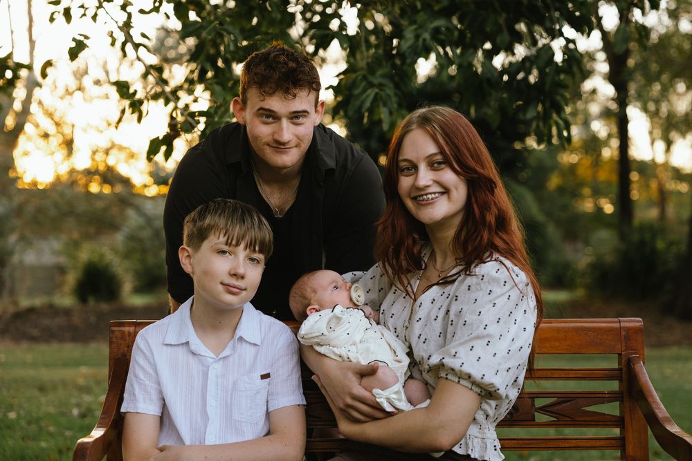 Family portrait of parents with teenage son and newborn baby sitting on a wooden bench in a sunlit park.