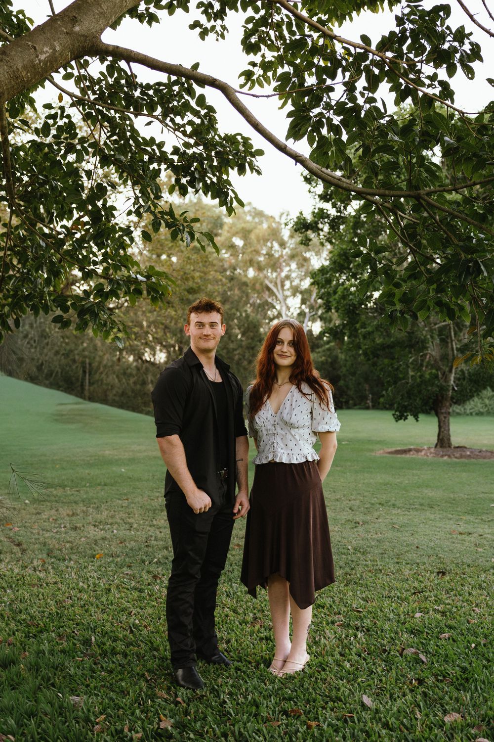 A couple stands hand-in-hand on green grass beneath a leafy tree, smiling at the camera in a park setting.