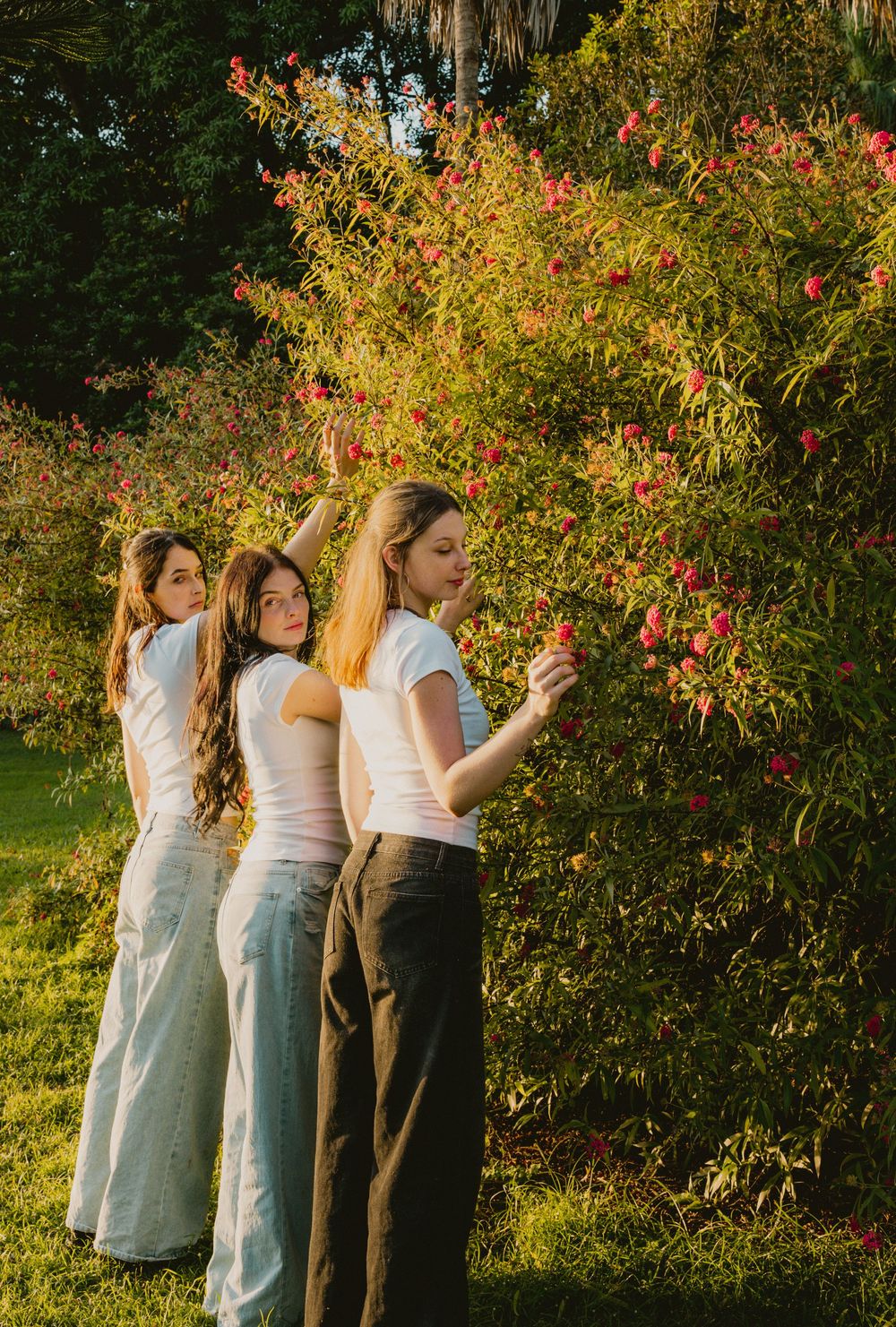 Three young women in white tops stand by a flowering bush in a sunlit garden.