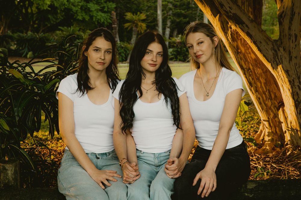 Three young women in matching white tops and jeans sit together outdoors near a tree in a lush green park.