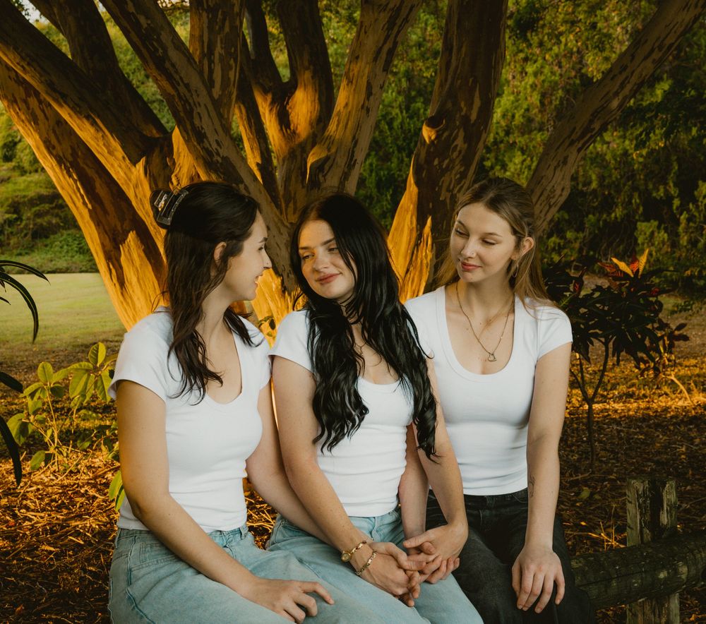 Three young women in white tops and jeans sit together outdoors near a tree in a lush green park setting.