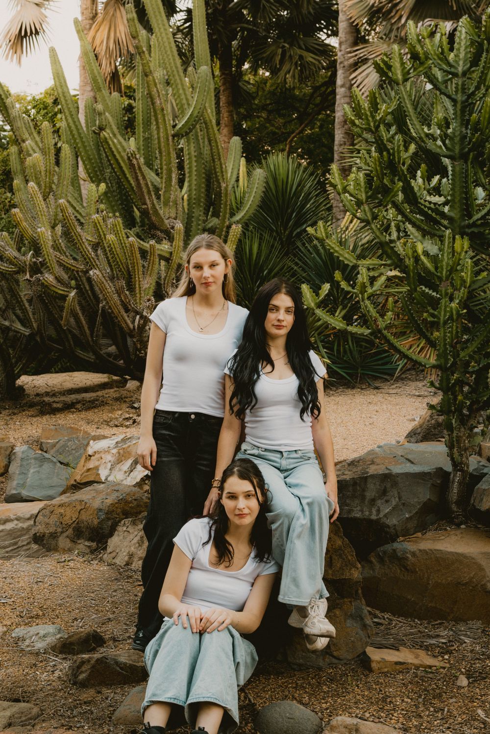 Three young women in white tops pose among tall cacti and rocks in a desert garden setting.