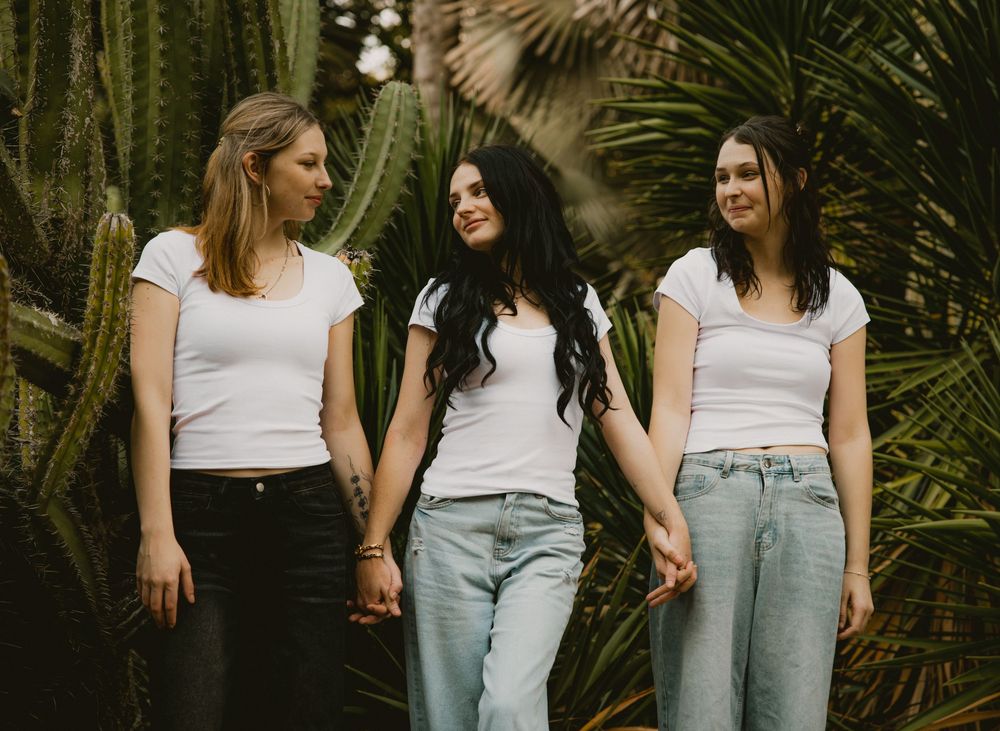 Three young women in white t-shirts and jeans hold hands among tall green cacti outdoors.