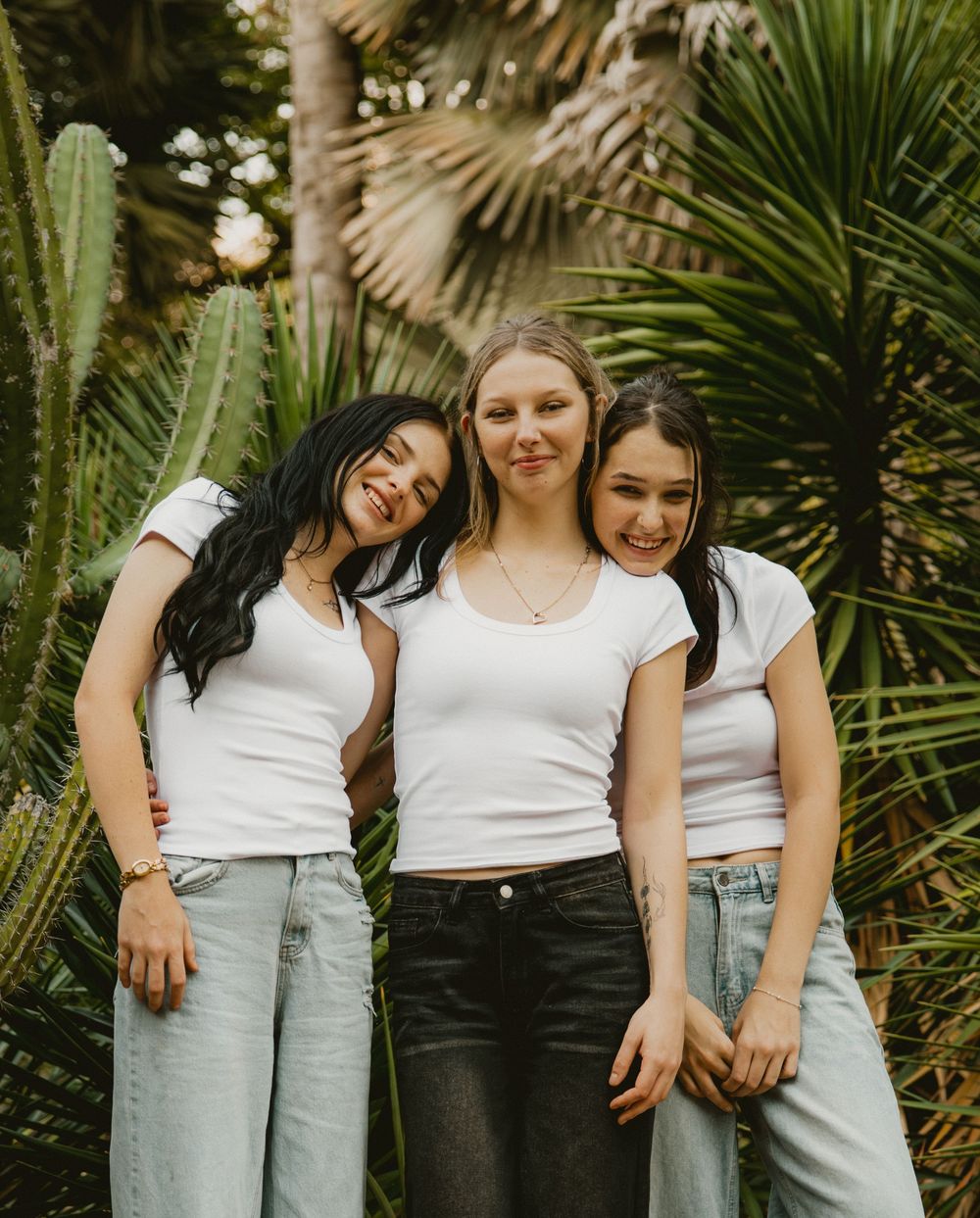 Three young women in white crop tops and jeans pose together smiling among tropical cacti and palm plants outdoors.
