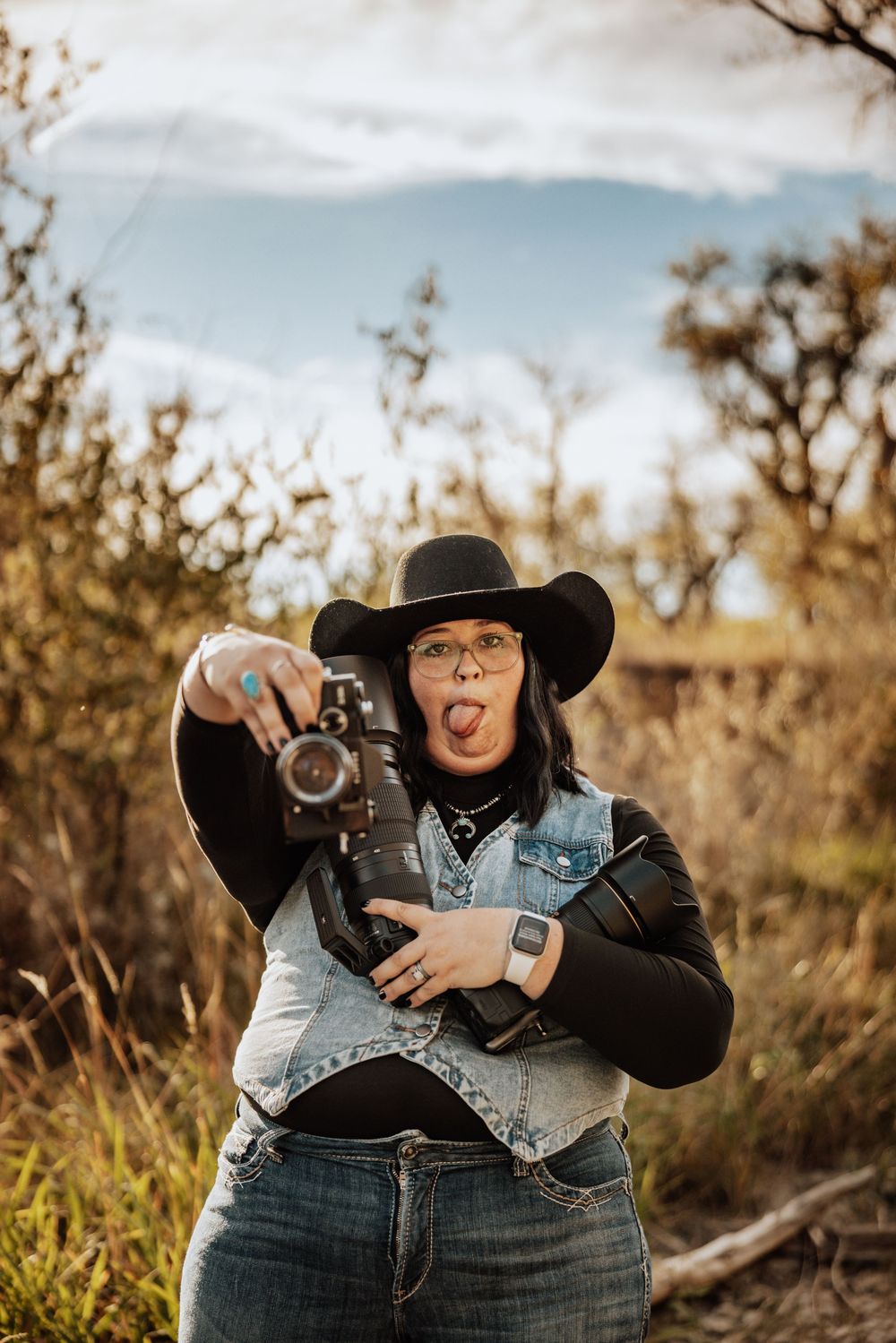 Woman in black cowboy hat holding two camera lenses on shoulders, smiling outdoors in a natural setting.