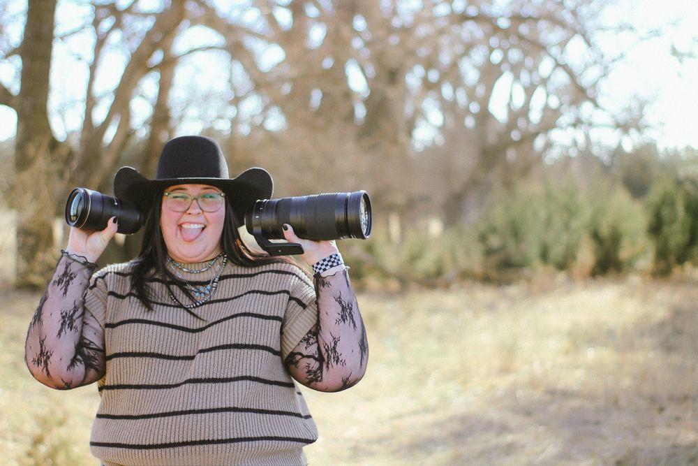 Woman in black cowboy hat holding two camera lenses on shoulders, smiling outdoors in a natural setting.