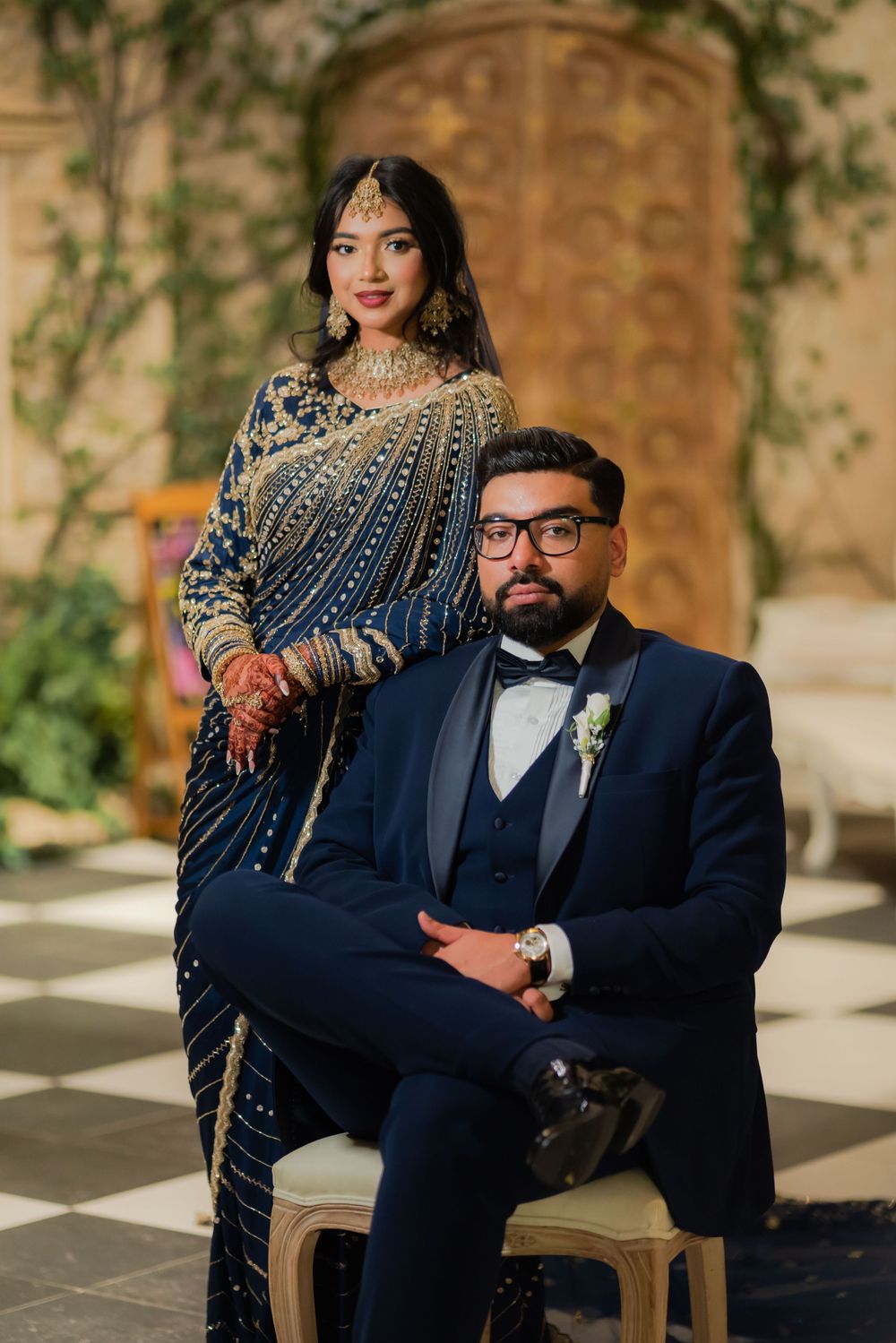 South Asian bride in navy blue embroidered saree stands beside groom in navy tuxedo seated on chair at wedding.