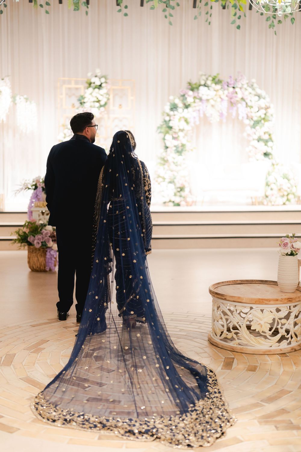 Bride in navy blue embroidered veil and gown walks with groom toward floral wedding altar decoration.