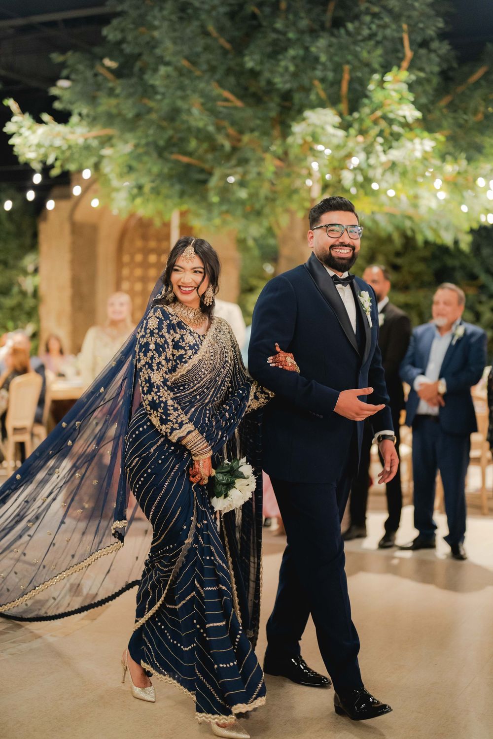 Bride in navy blue embroidered saree and groom in navy suit smile while walking at their elegant wedding reception.
