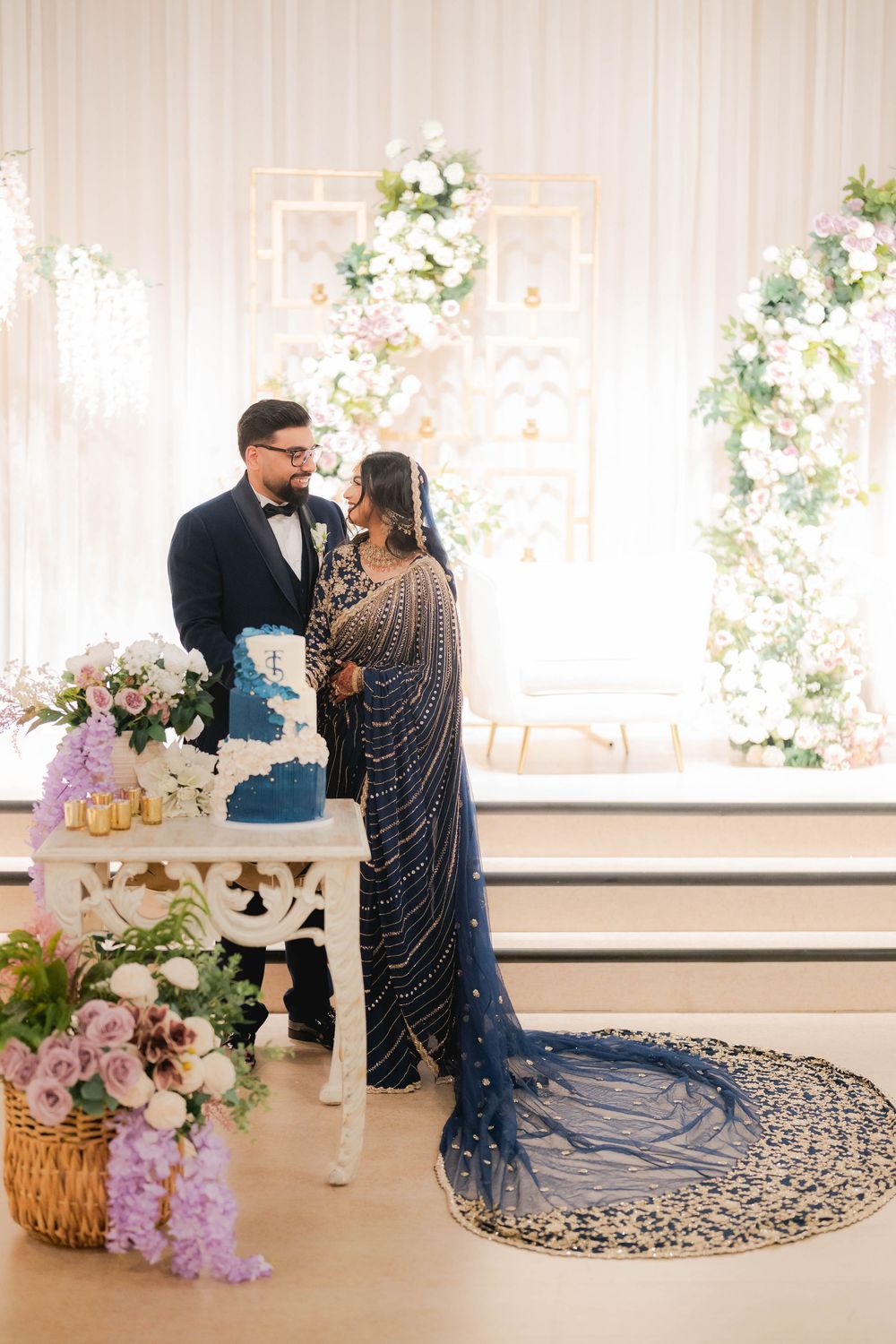 Bride in navy beaded saree and groom in tuxedo stand together at blue wedding cake table with floral decor.