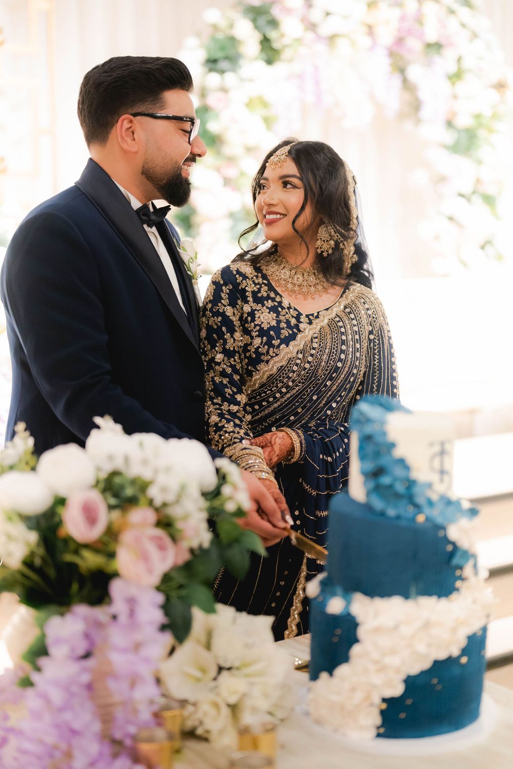 Smiling South Asian bride in embroidered navy saree holds groom's hand beside elegant blue tiered wedding cake.