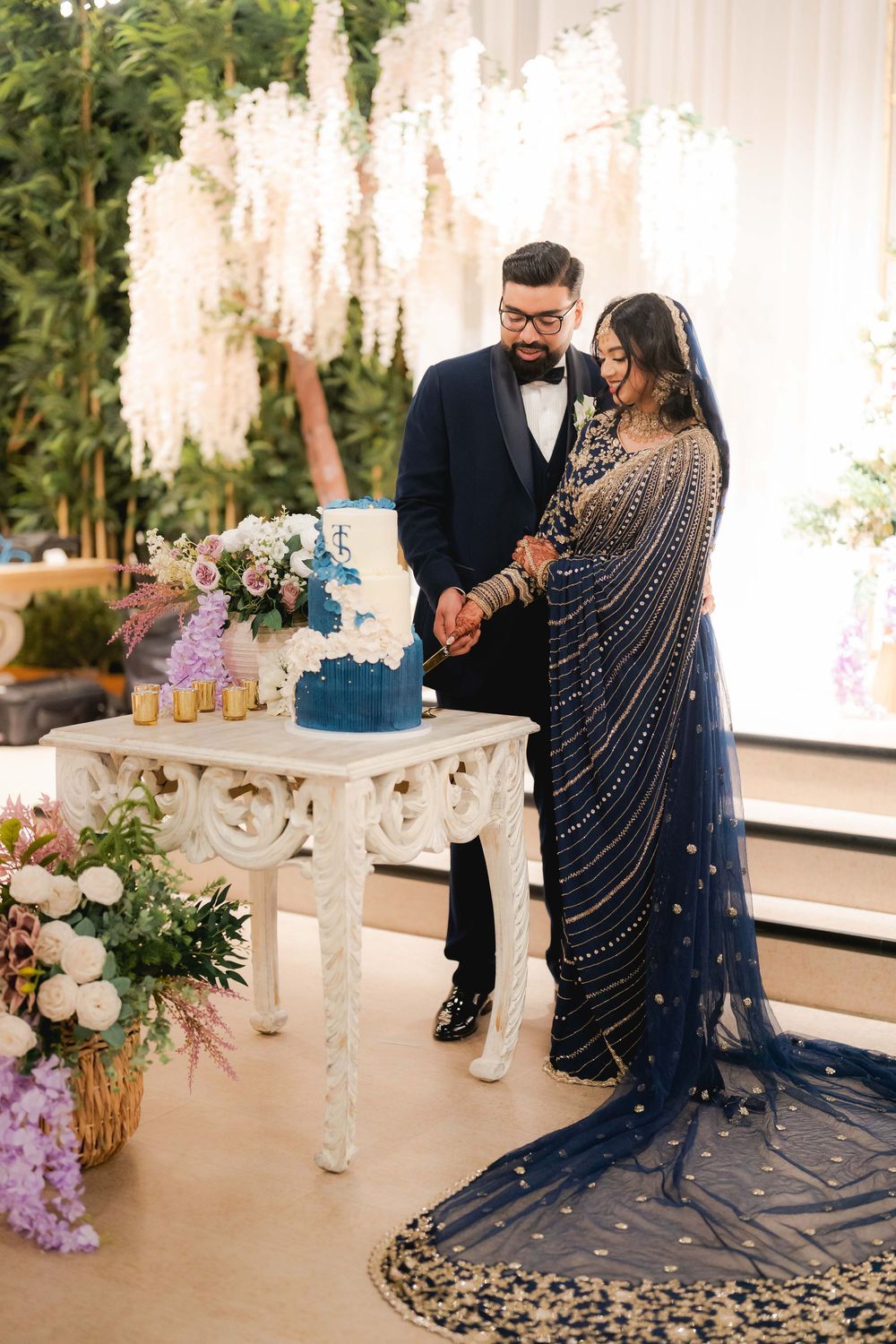 South Asian couple in matching navy outfits cutting blue wedding cake at elegant floral reception.