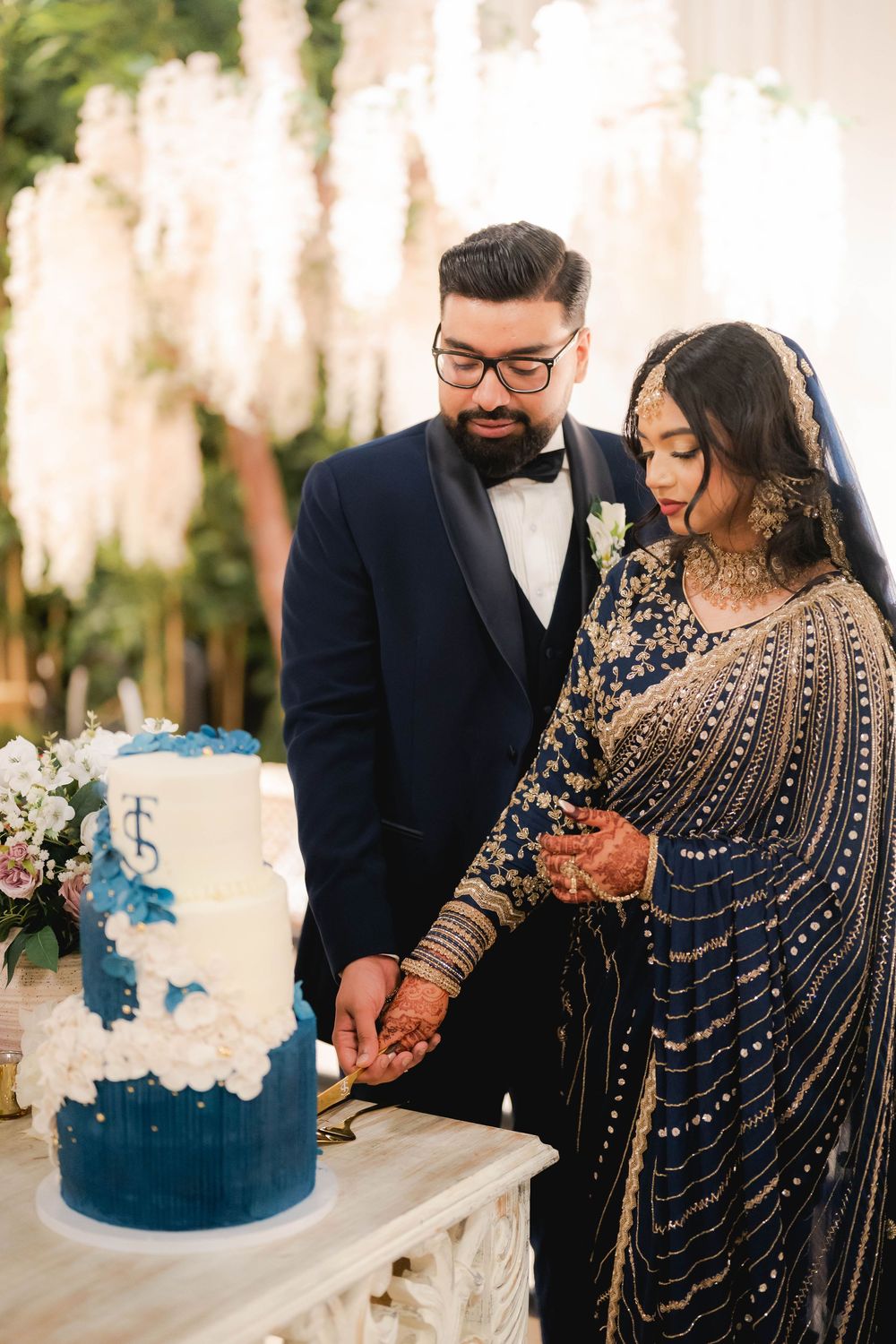 Bride in navy embroidered saree and groom in tuxedo cutting elegant blue and white tiered wedding cake.