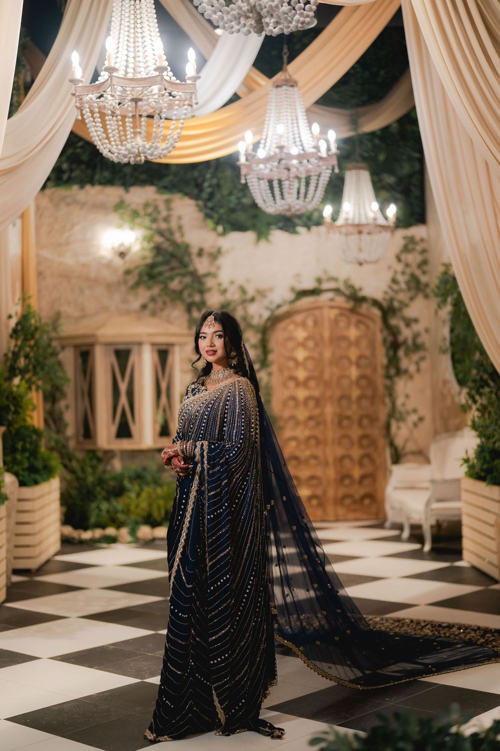 Woman in navy blue embellished saree poses under crystal chandeliers in an elegant decorated venue.