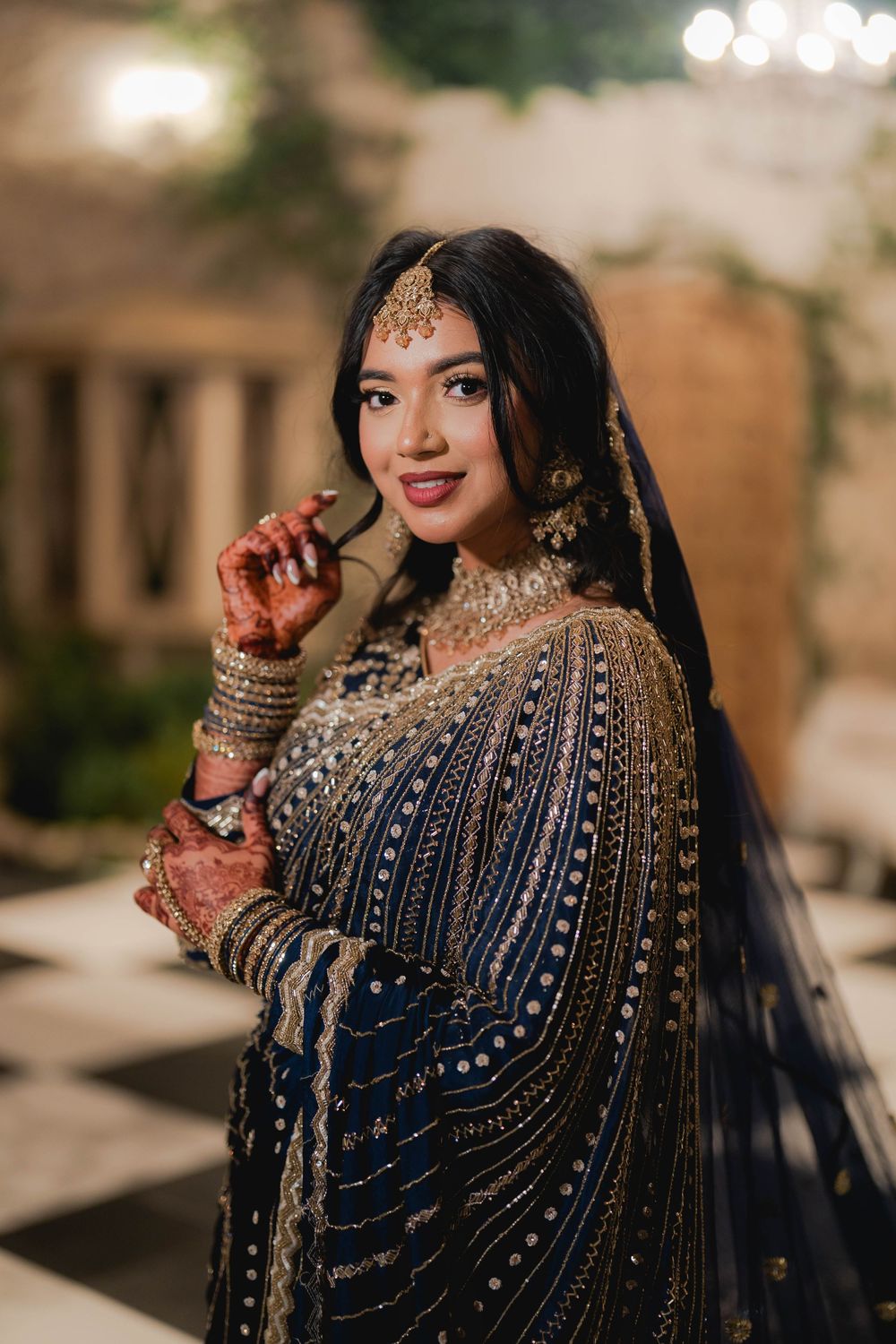 Bride in navy blue embroidered lehenga with gold jewelry and mehndi poses elegantly at an Indian wedding.