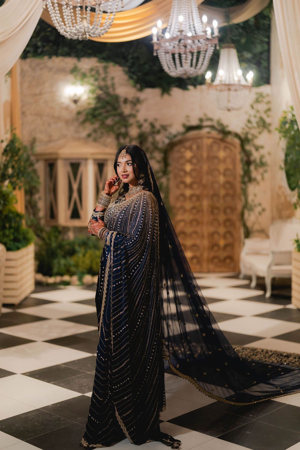 Bride in navy blue embellished saree with dupatta posing in elegant venue with crystal chandeliers.