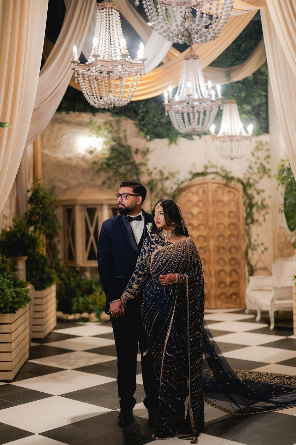 Couple in matching navy outfits posing under crystal chandeliers in elegant black and white venue.