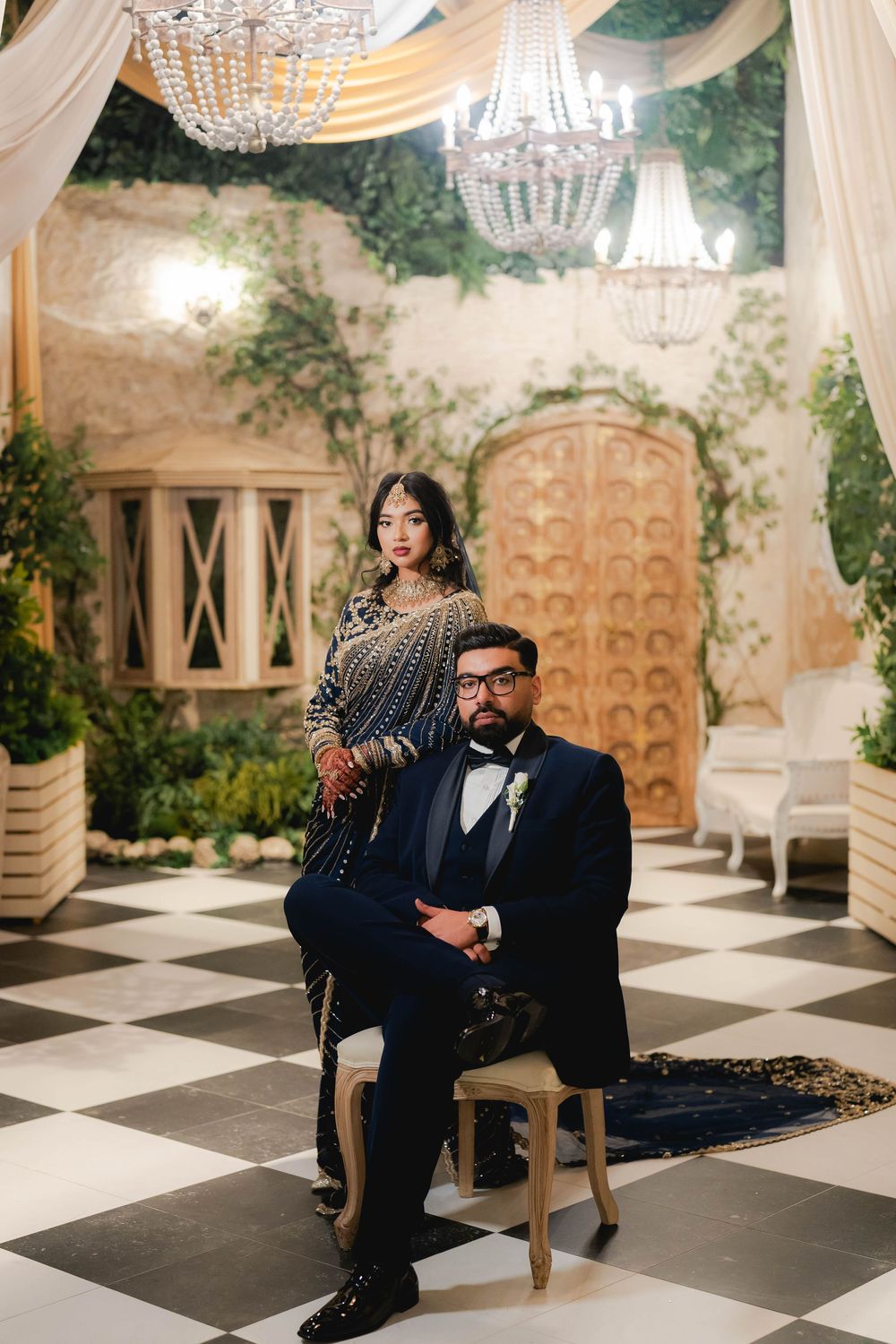 Elegant couple in Indian wedding attire pose in a grand venue with chandeliers and checkered floors.
