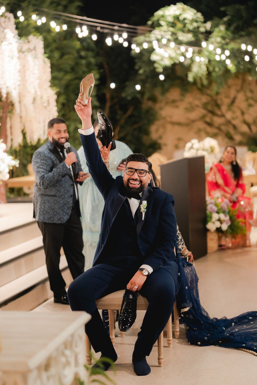 Groom in navy suit joyfully raises a shoe during a fun wedding game, with guests and festive lights in background.