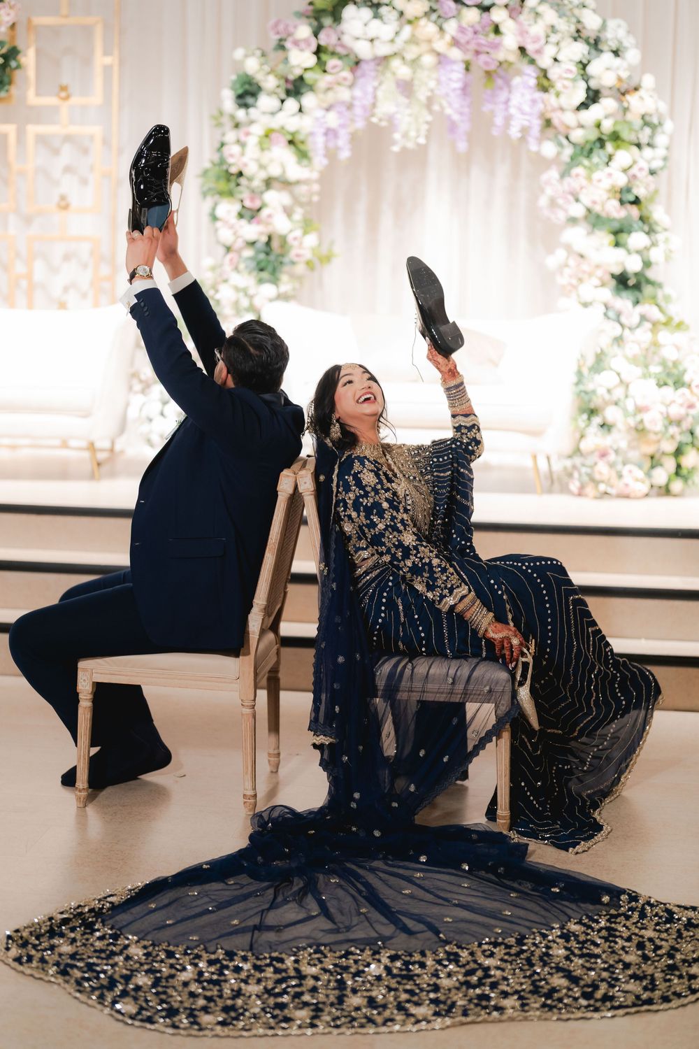Bride and groom in navy outfits playfully hold up shoes while seated at their elegant floral wedding reception.