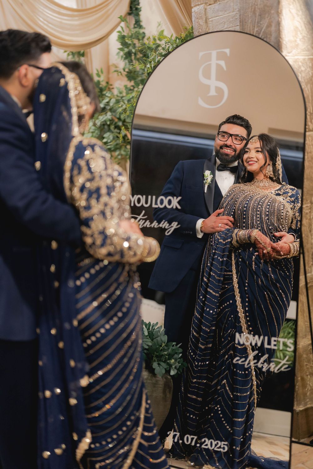 South Asian bride and groom in navy blue embroidered wedding attire pose by decorative mirror at reception.