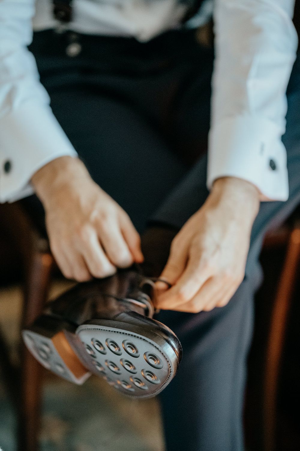 Man in formal attire tying brown dress shoes, wearing white shirt and dark suspenders.