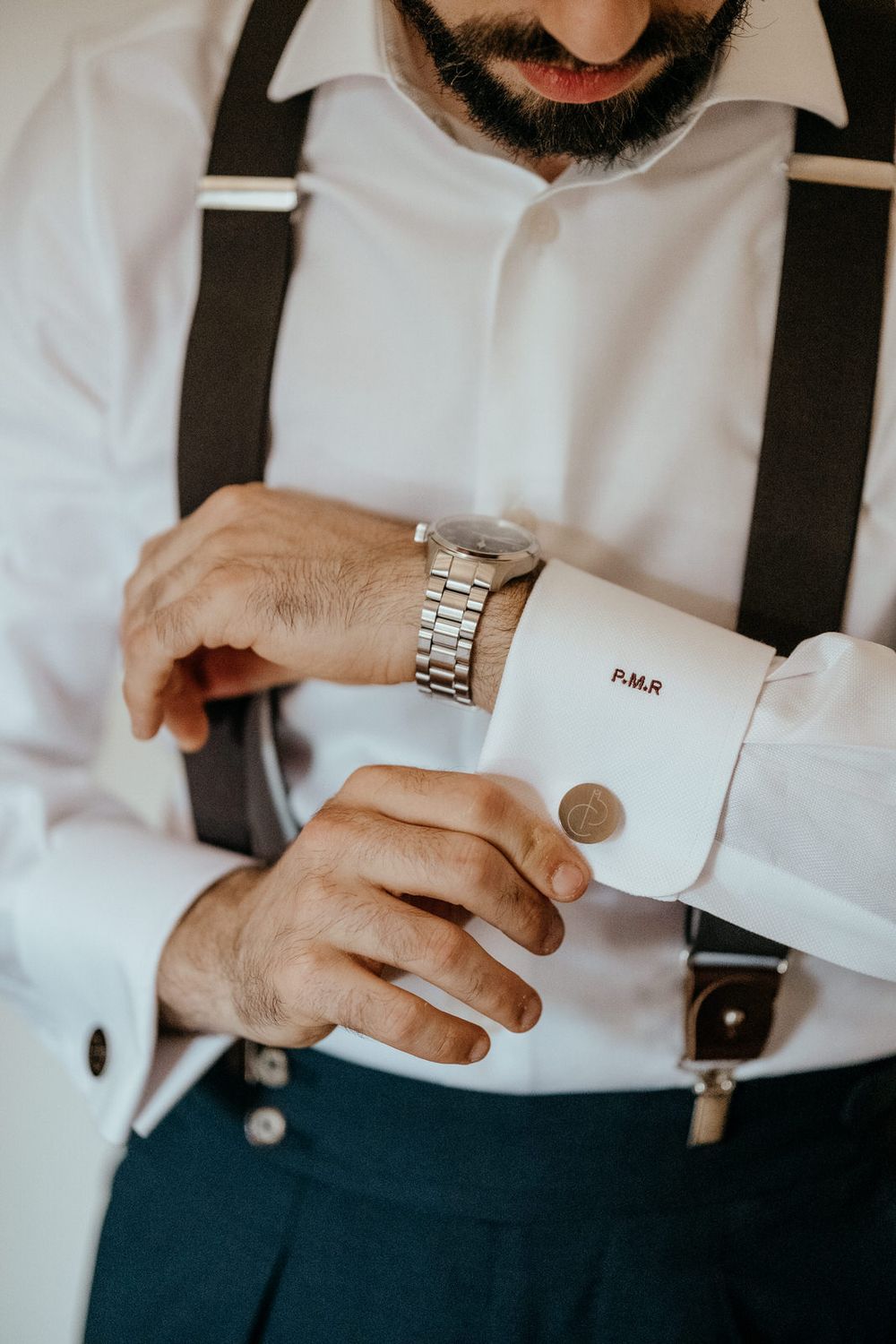 A bearded groom in a white dress shirt with monogrammed cuffs, suspenders, and a silver watch adjusts his cufflinks.
