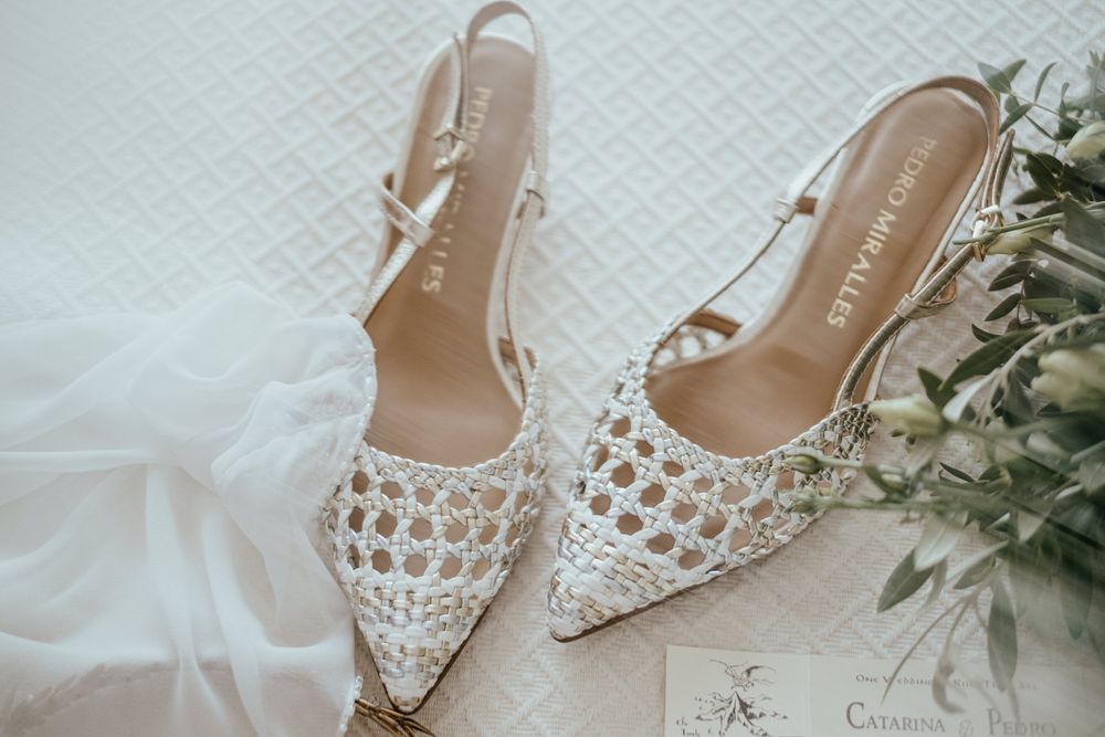 Elegant white woven slingback bridal heels displayed beside a wedding dress and floral bouquet.
