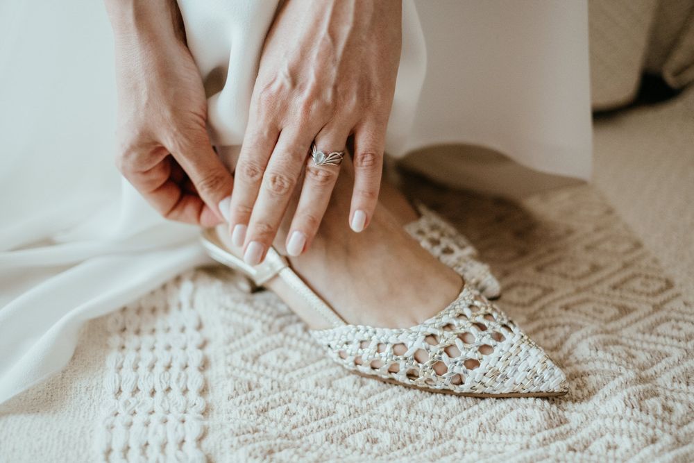 Bride wearing a diamond ring fastening a strap on white woven pointed-toe wedding flats.