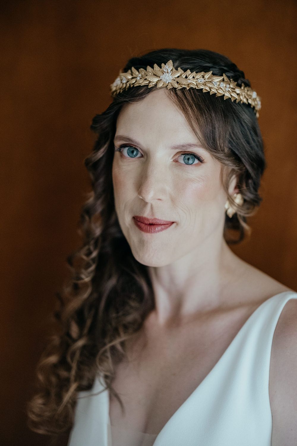 A bride with blue eyes wearing a gold floral crown headpiece and white dress, with curled brown hair.