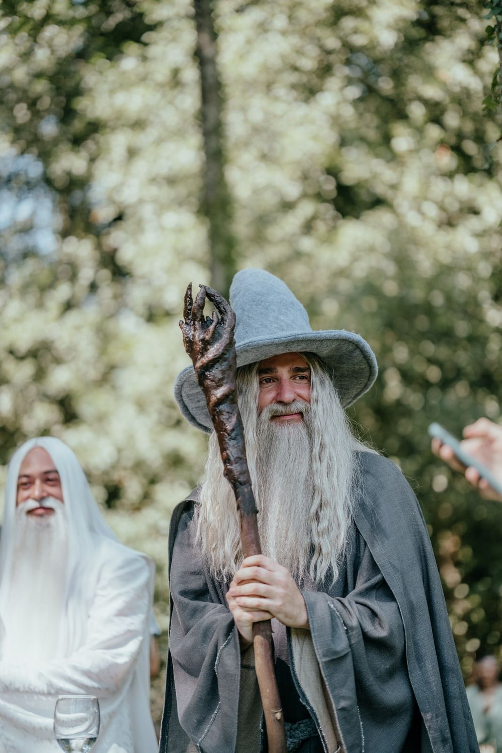 Man dressed as Gandalf wizard with gray robes, long white beard, and wooden staff at outdoor cosplay event.