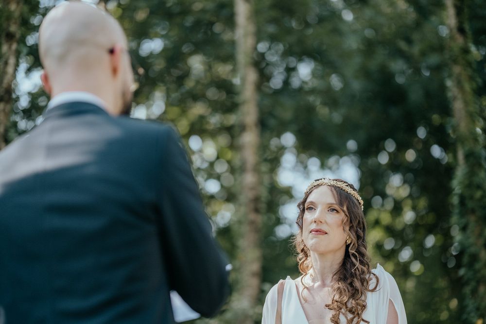 Bride gazing lovingly at groom during outdoor wedding ceremony surrounded by lush green trees.