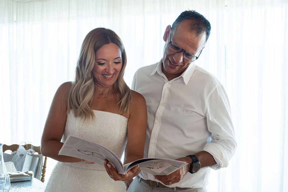 A smiling bride and groom in wedding attire reading a book together at their reception venue.