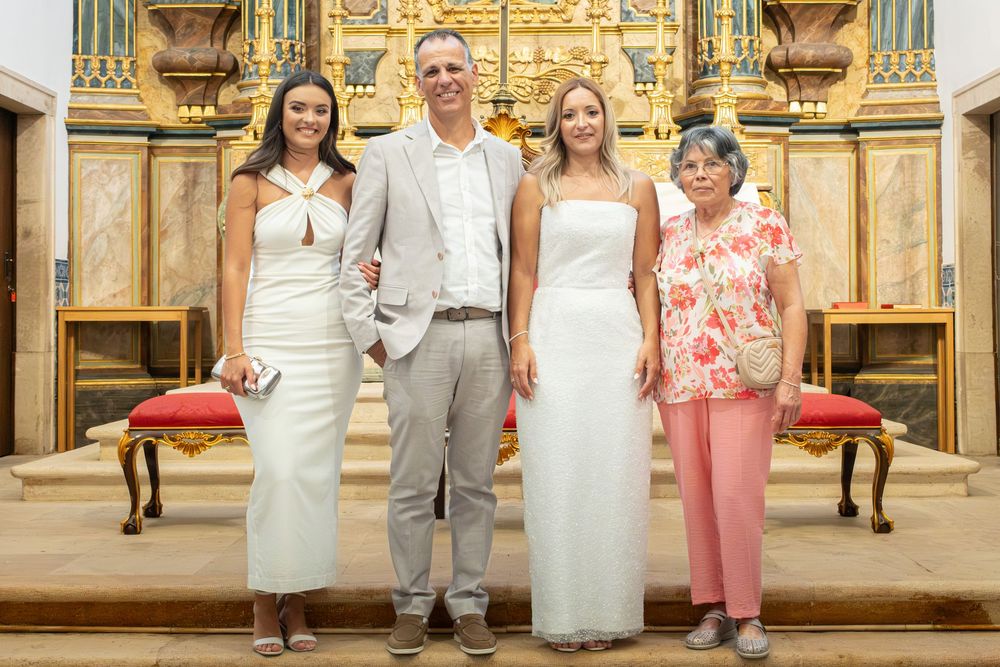 Four people posing together in a church, two women in white dresses, a man in gray suit, and an older woman in pink.