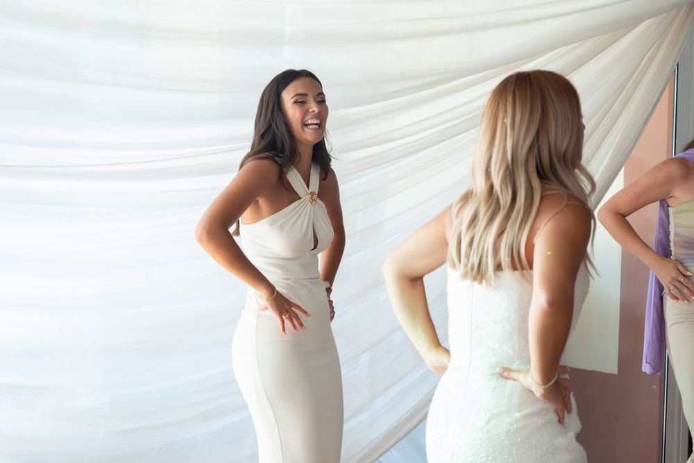 Two women in elegant white dresses laughing together at a wedding fitting or bridal event.