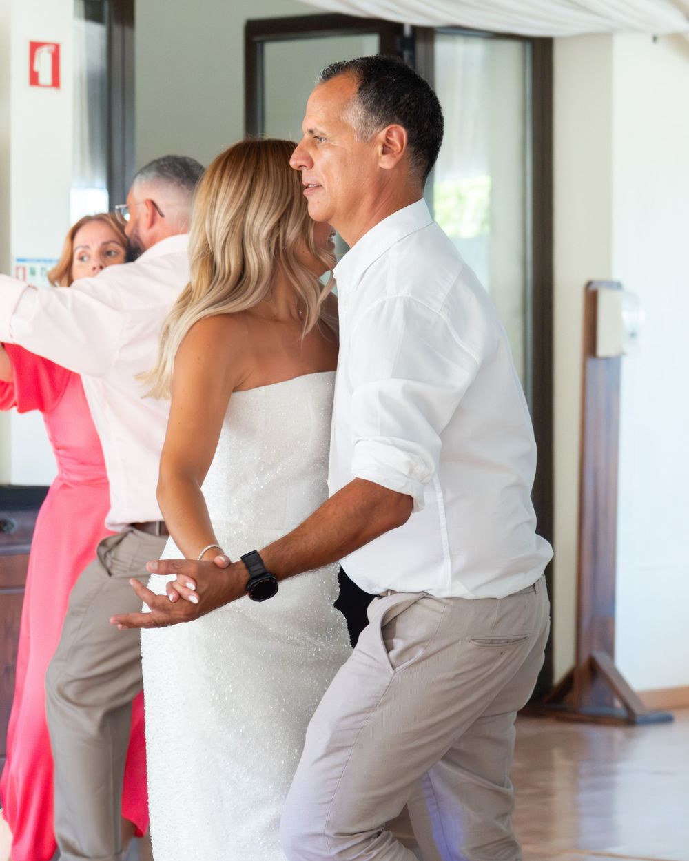 Bride and groom share a dance at an outdoor wedding reception, both dressed in white, with guests visible in the background.